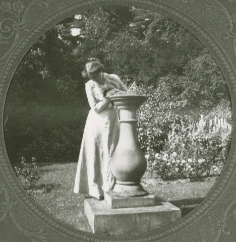 White woman in a long dress leaning on a large urn in a garden.