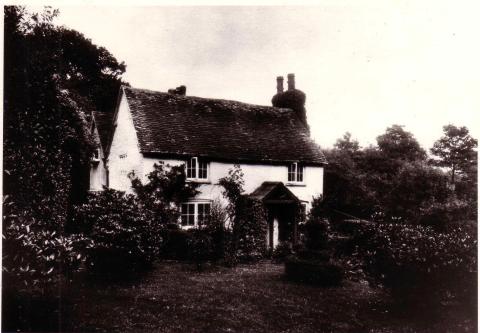 Black and white image of two-storey, white brick cottage amidst trees and garden.
